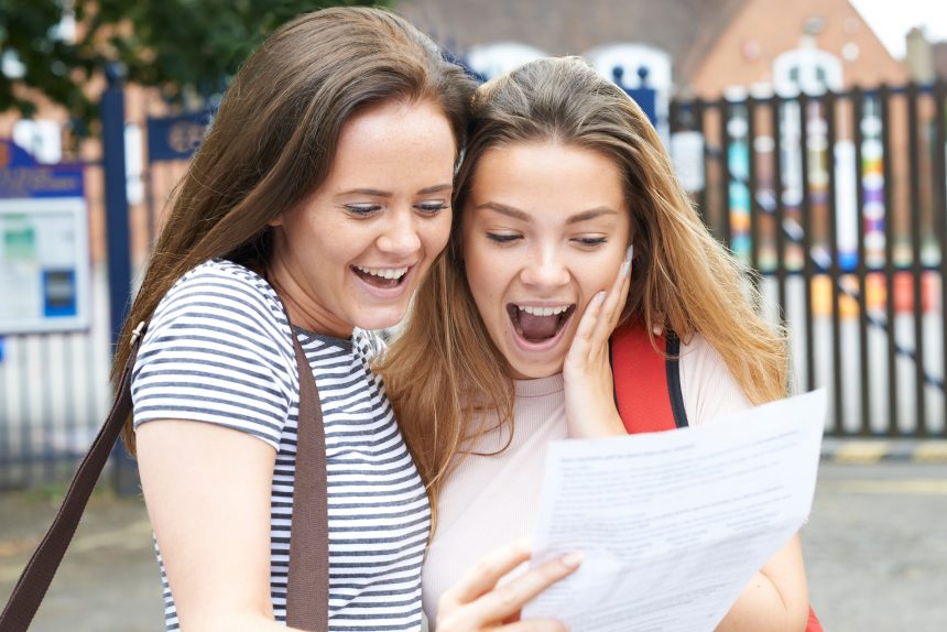 A happy and surprised teenage girl looking at her exam results on A level Results Day with her friend.