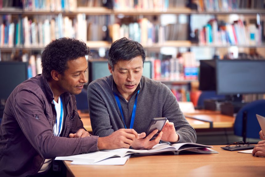 Two teachers sitting at a table in their school library and looking at a mobile phone.