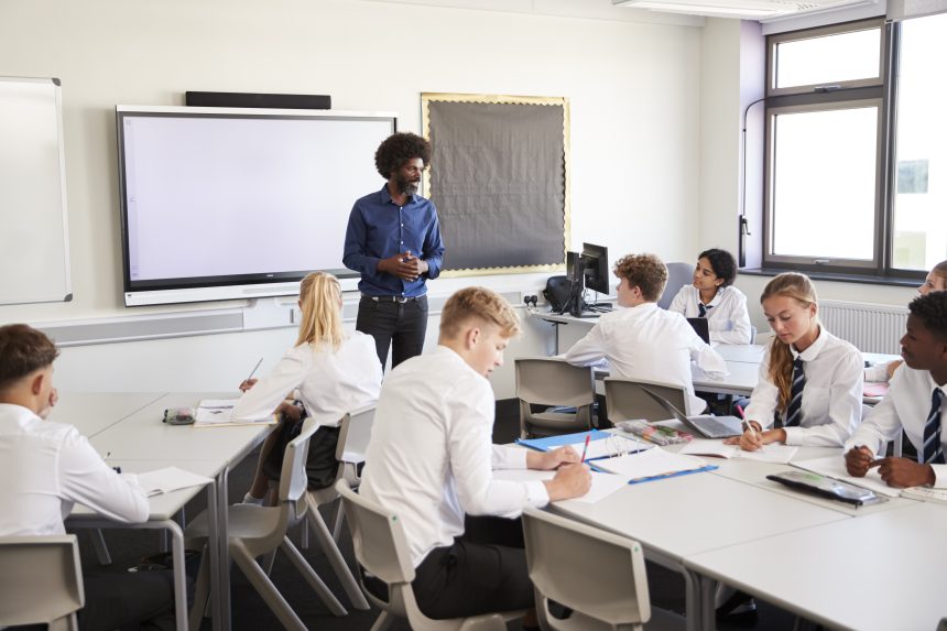 A male secondary school teacher standing next to an interactive whiteboard and teaching a lesson to his pupils.