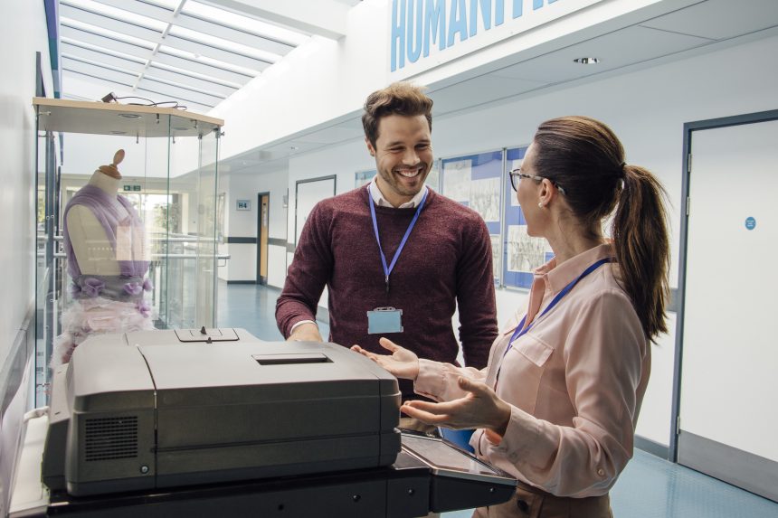 Two teachers chatting and catching up while standing at a printer in the school corridor.