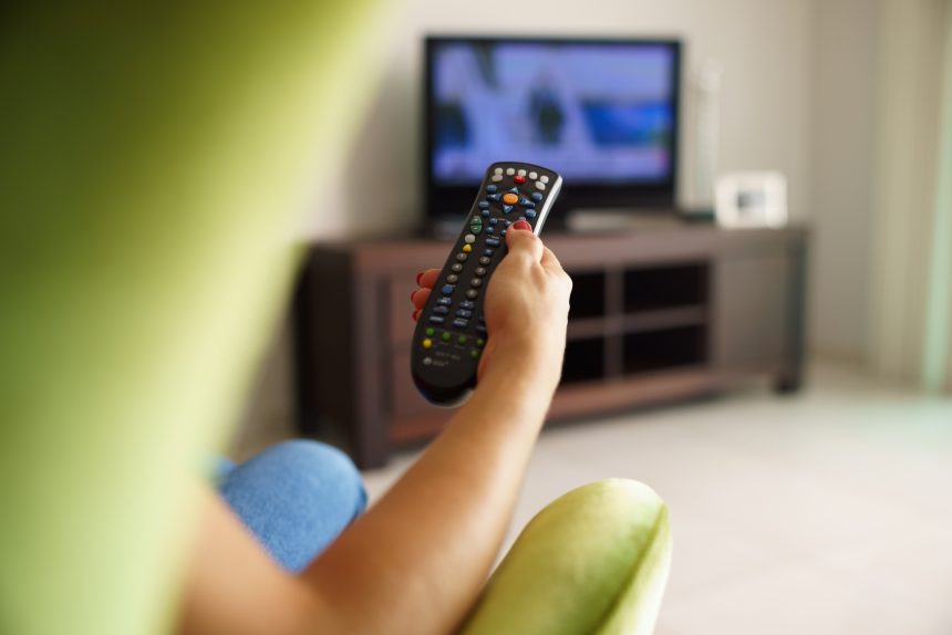 Over-the-shoulder view of a woman holding a TV remote and surfing for television programmes.