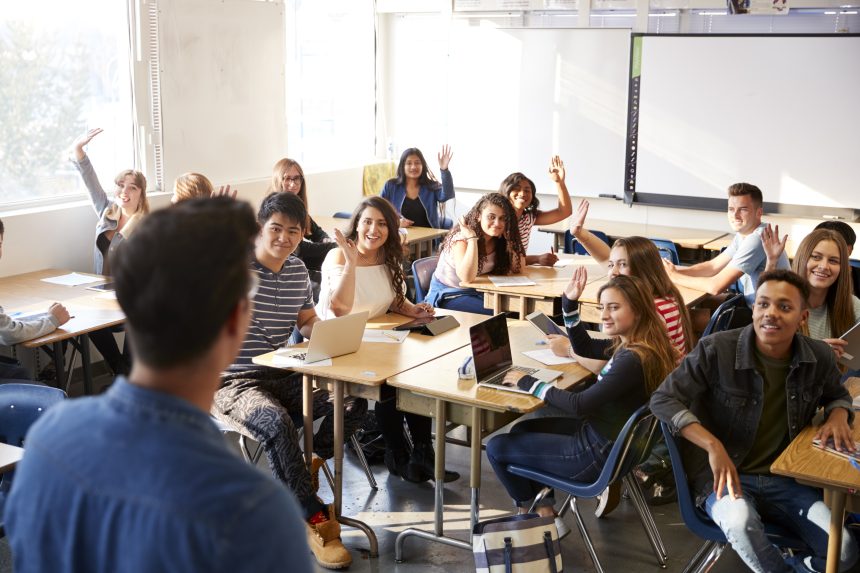 A group of students in a classroom. A teacher is stood at the front and some students have their hands raised.