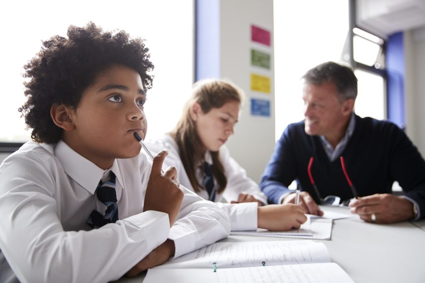 A male student thinking whilst a teacher talks to a pupil in the background.