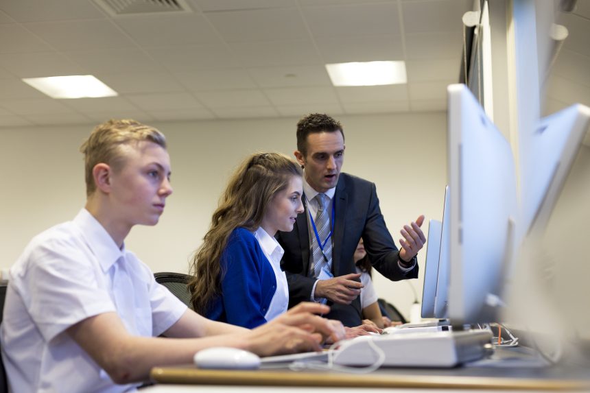 Teacher giving feedback to two students sat at computers.