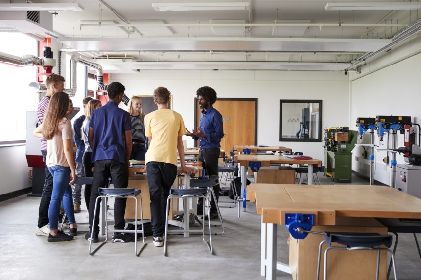 Group of A Level students standing around a workbench and listening to their teacher in product design class.