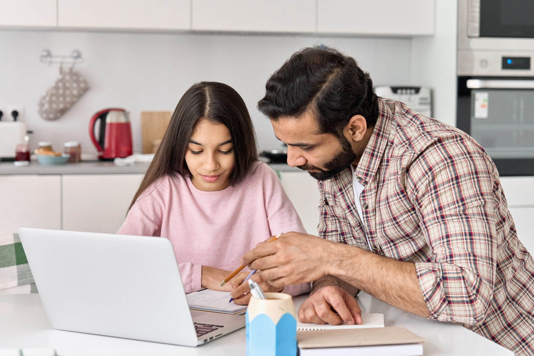 A dad is helping his daughter with her exam revision as they sit at the kitchen table surrounded by a laptop, notebook, and pens.