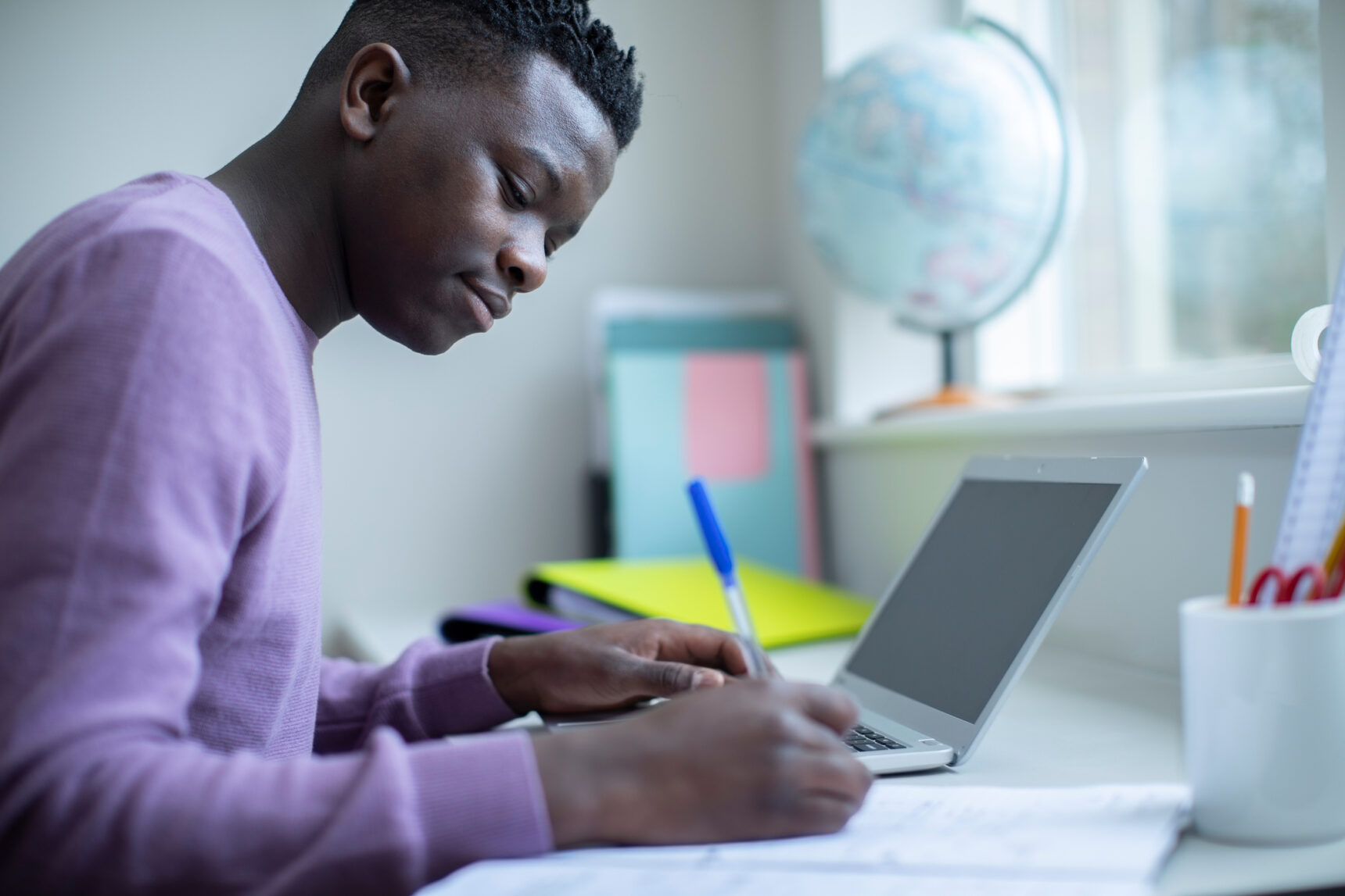 A teenage boy is writing notes at a desk as he studies for exams surrounded by folders, a laptop, and lots of natural light from a window nearby.