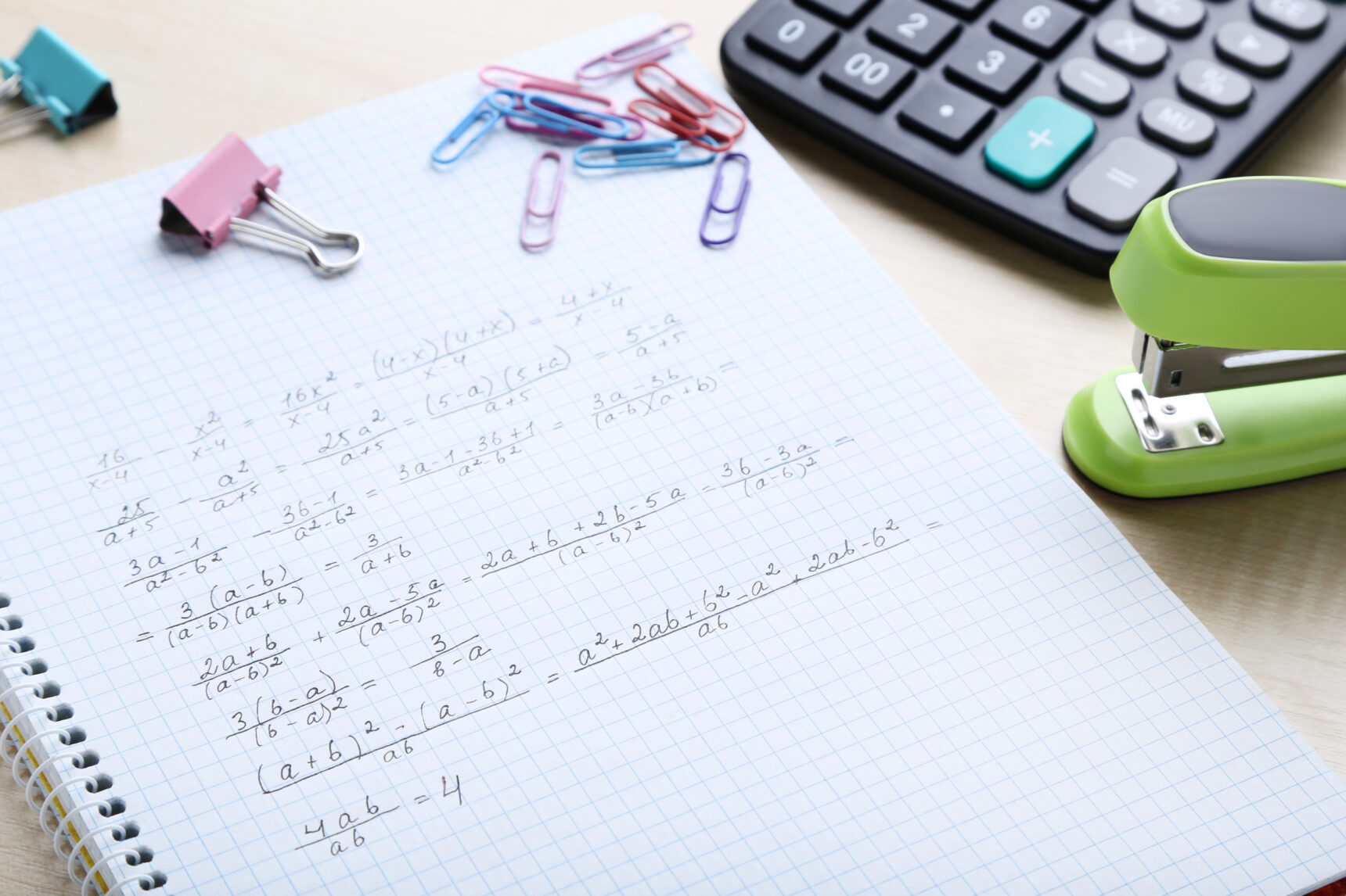 A maths notebook showing algebra on squared paper next to a calculator and stapler.