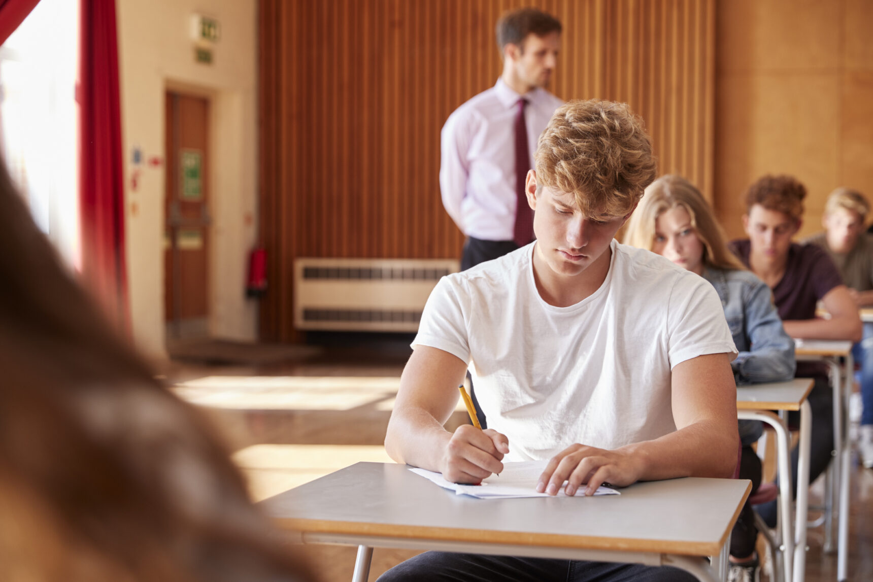 A teenage boy is sitting a GCSE exam in an exam hall.