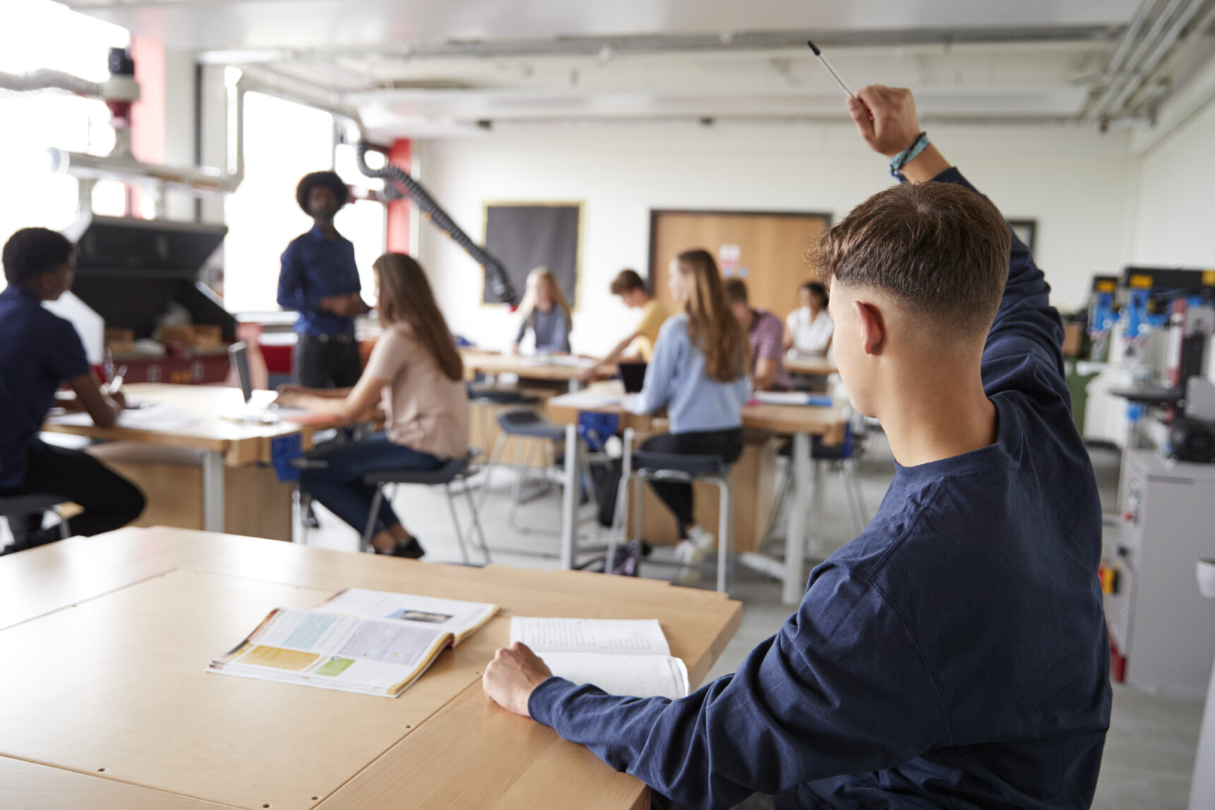 A student raises their hand to ask a question while a teacher speaks to the class.