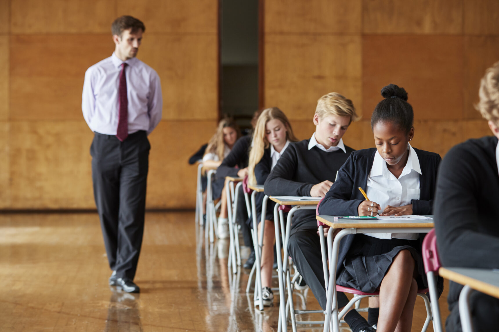 Students sitting AQA GCSE English Language Paper 1 in an exam hall with a teacher invigilating.