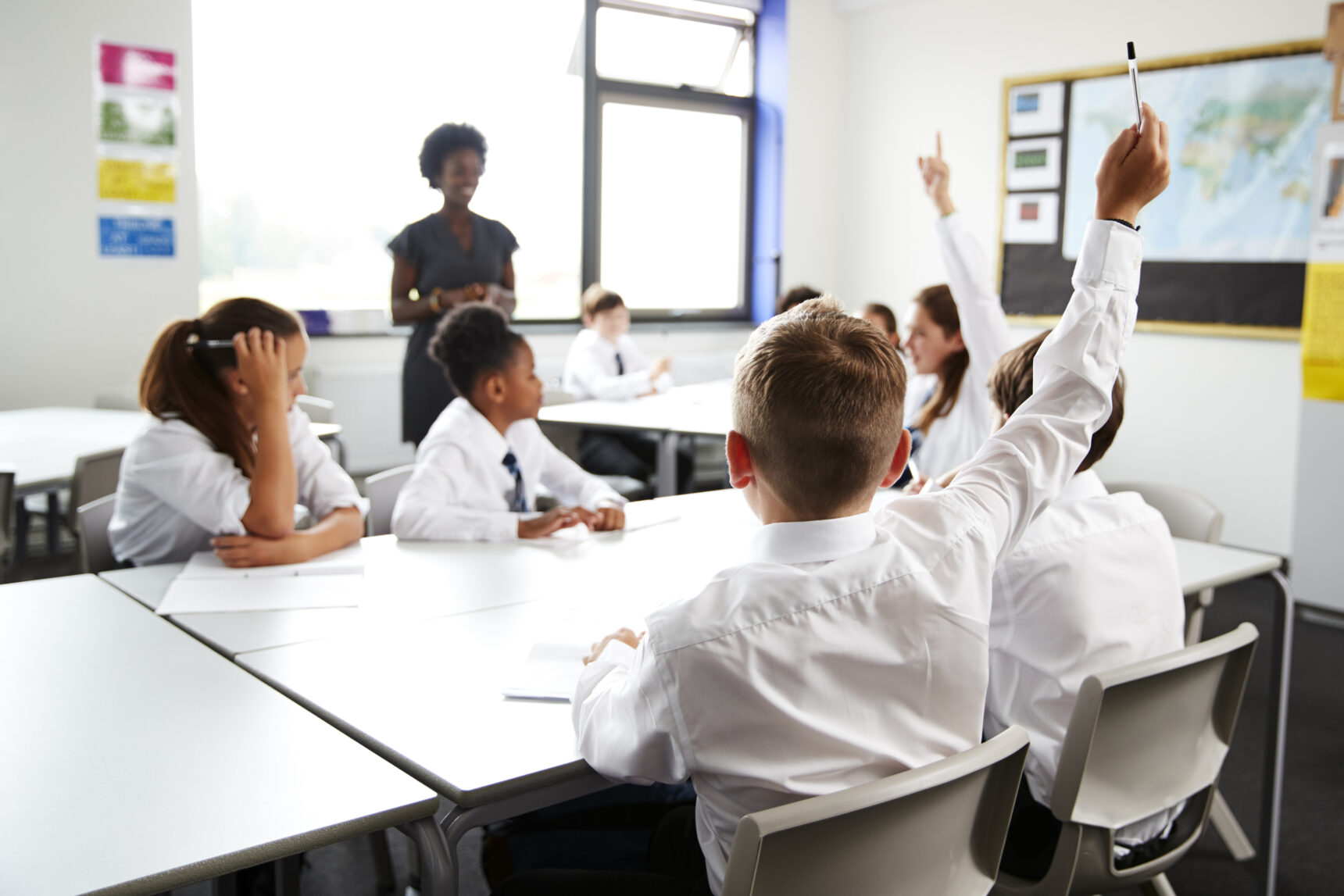 Young studetns in a classroom are raising their hands to answer a hinge question from the teacher.