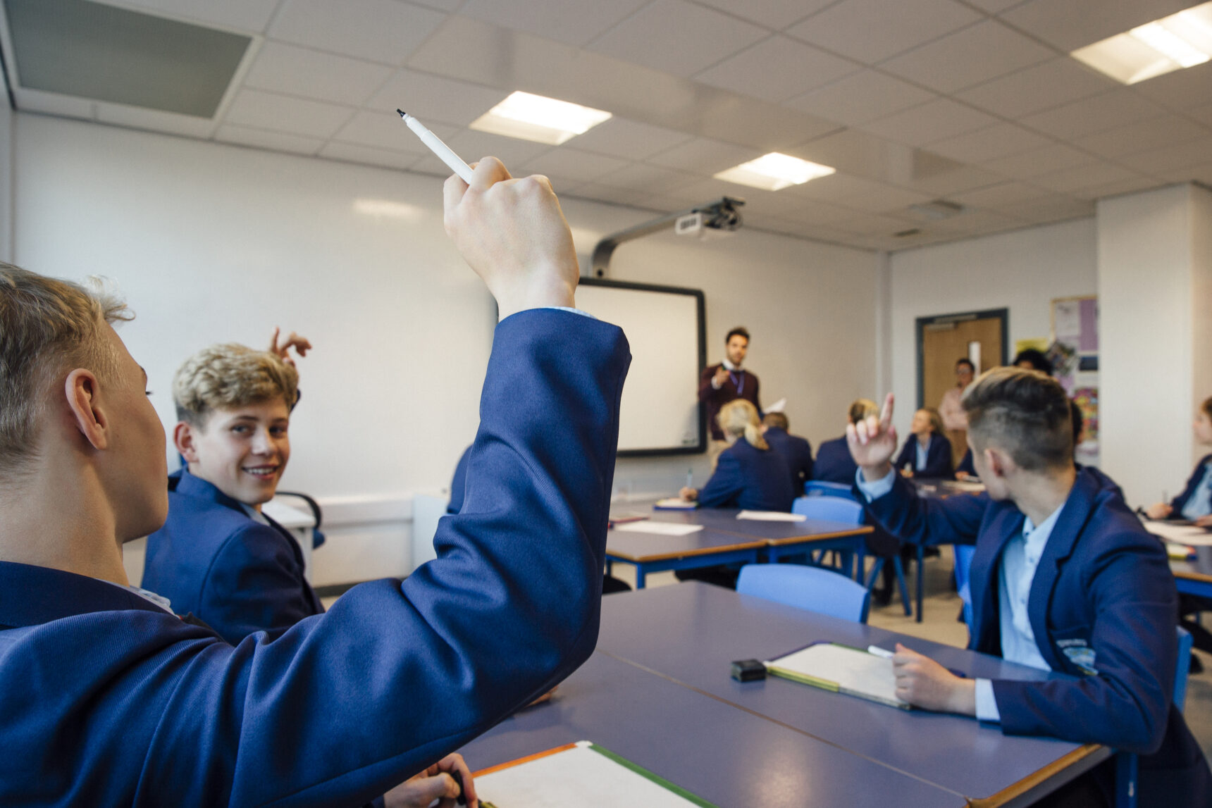 Students in a classroom are smiling and raising their hand to answer a hinge question that the teacher has presented during a lesson.