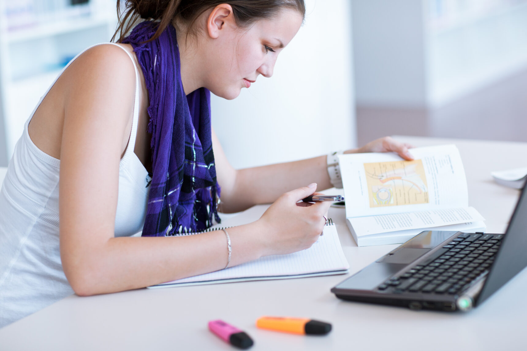 A teenage girl is taking notes from a textbook as she studies for exams.
