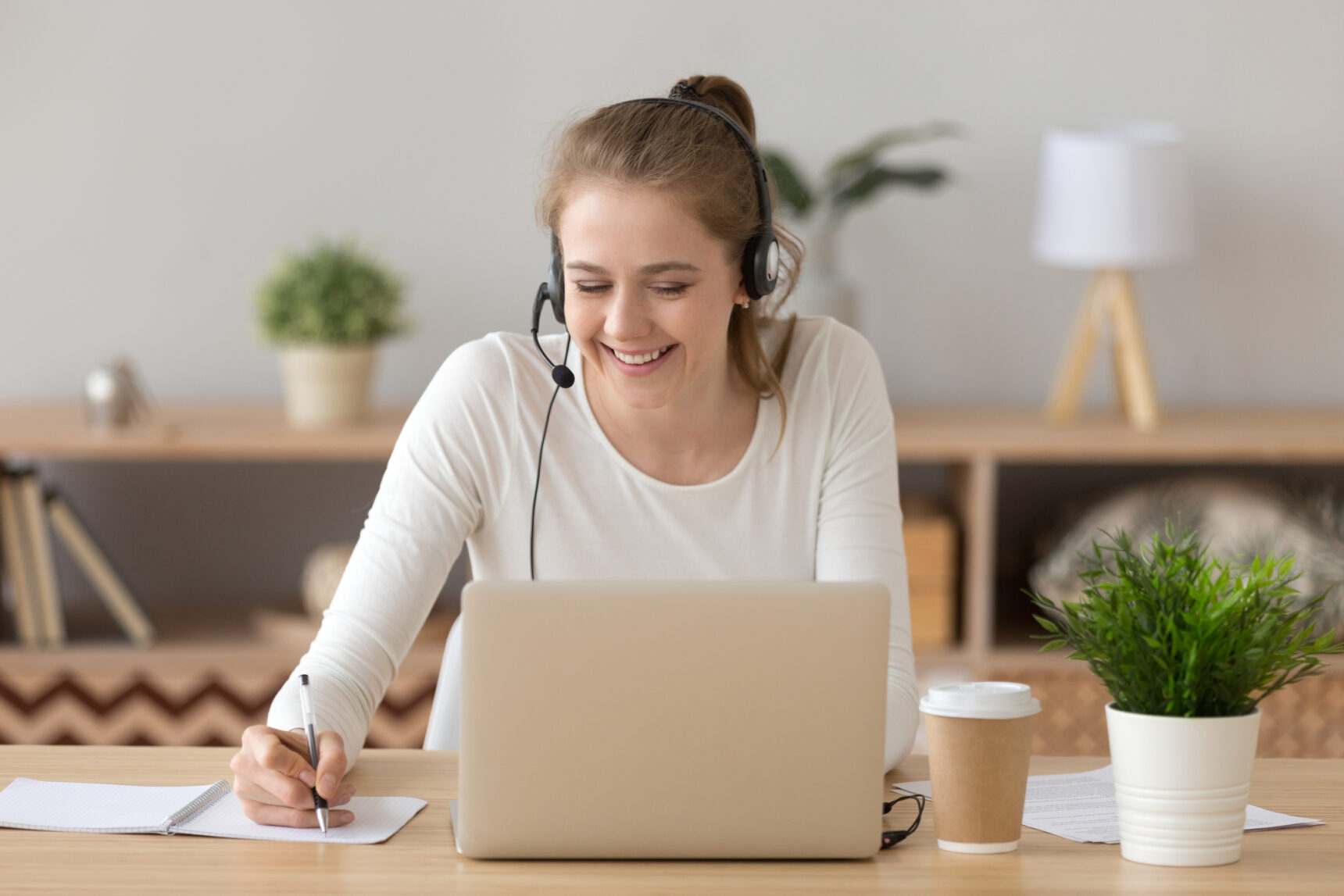 A female online tutor is smiling and writing notes as she teaches an online lesson.