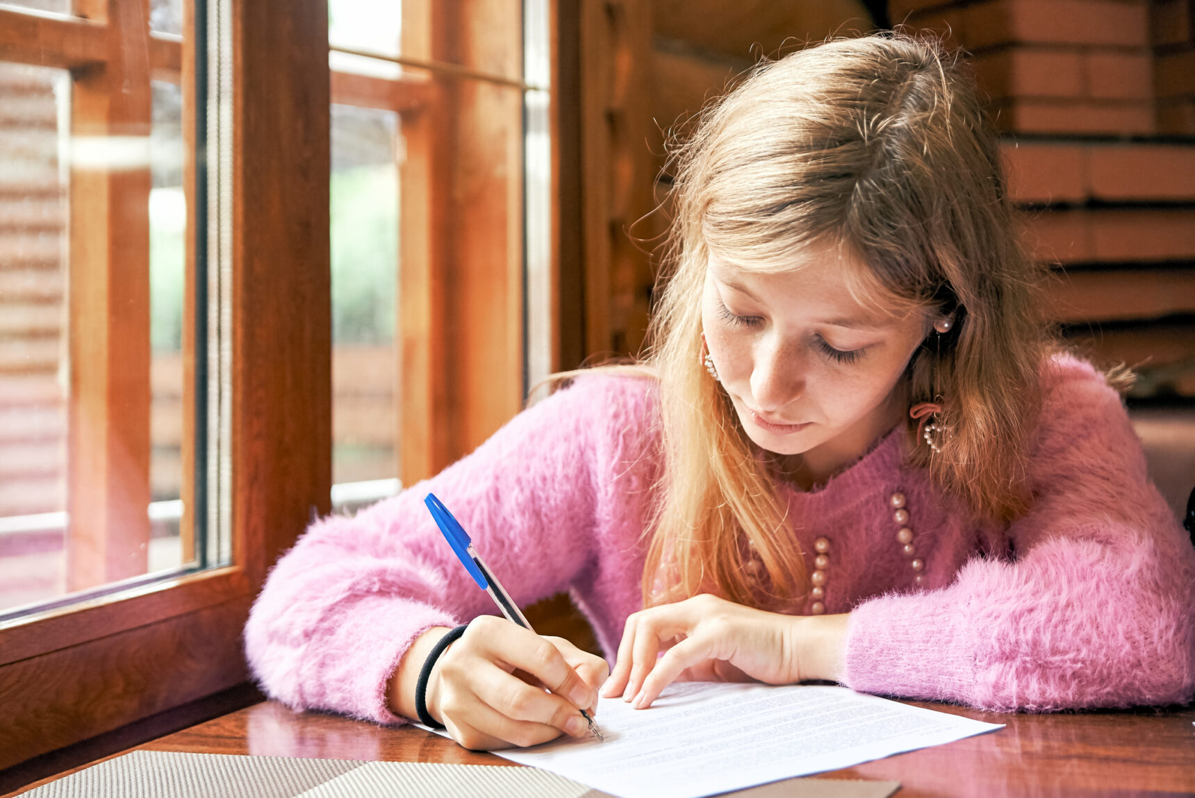 A teennage girl sitting near a window is practising writing for her GCSE English Language exam.