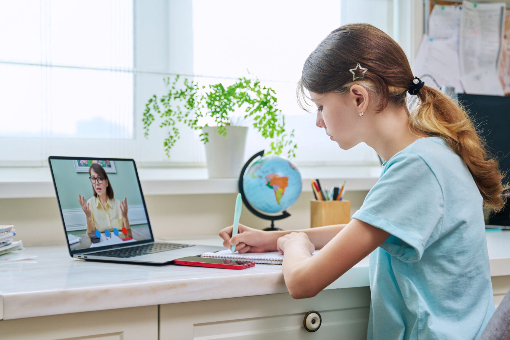 A young girl is sitting at a desk and taking notes during an online tutoring session.
