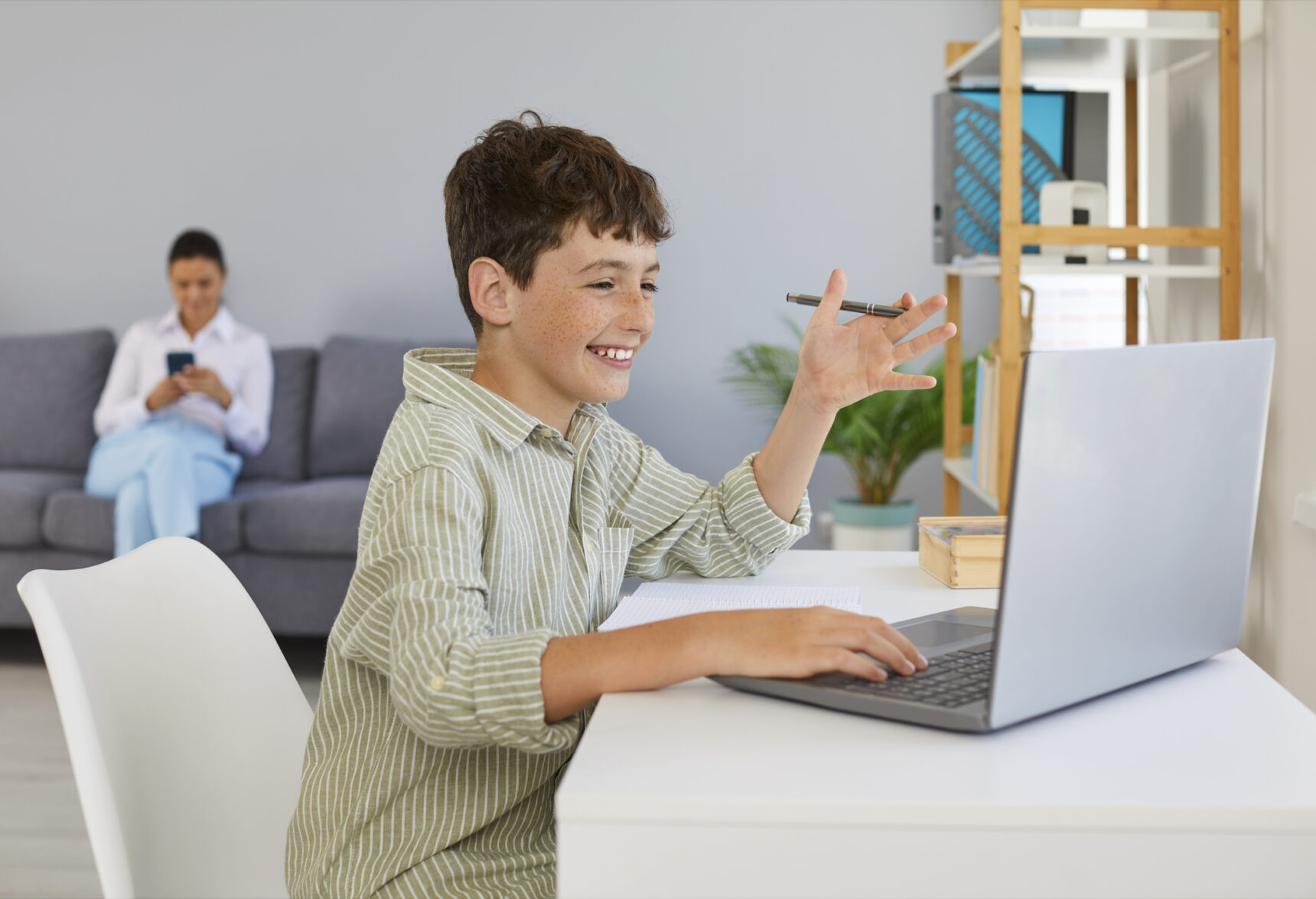 A young boy is chatting to his online tutor while his mother sits quietly looking at her phone in the background.