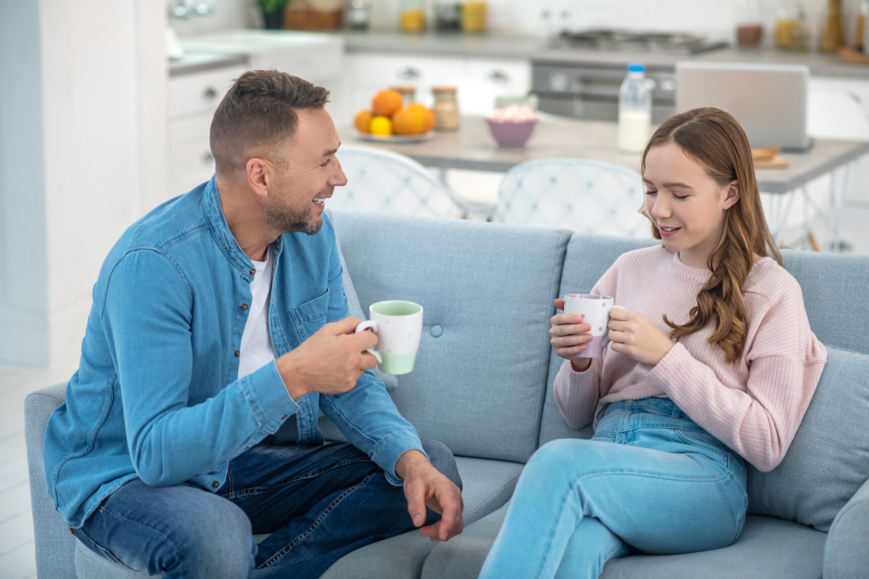 A dad and daugther are sitting on the sofa drinking tea and chatting.