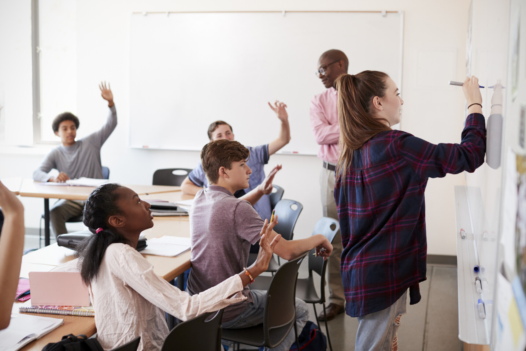 A class of students raise their hands to answer a question as one student writes on the whiteboard.