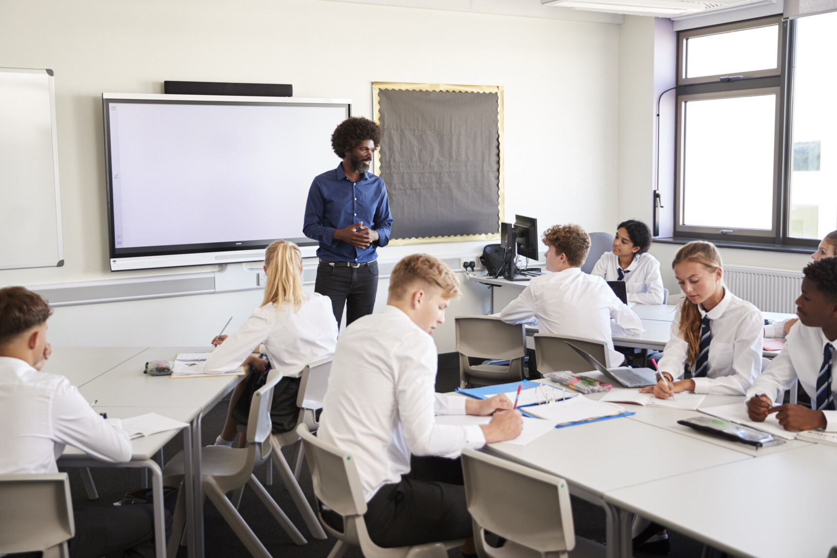 Students wrie notes during a lesson as the teacher speaks next to the whiteboard.